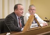 Representative Huot sits at the testifiers' table and speaks to someone out of view. Brad Johnson sits next to him and watches as he speaks.